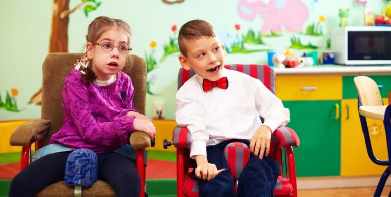 Foto de duas crianças com deficiência - uma menina e um menino - em cadeira de rodas coloridas. Elas estão em uma escolinha infantil