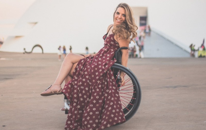 Carla Maia posa na cadeira de rodas com as pernas cruzadas. Ela está em frente ao Museu Nacional de Brasília