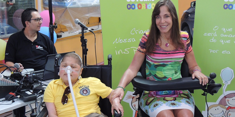 Foto da Mara com a mão sobre o braço do Adolfo Luis que utiliza respirador. Ambos em suas cadeiras de rodas.