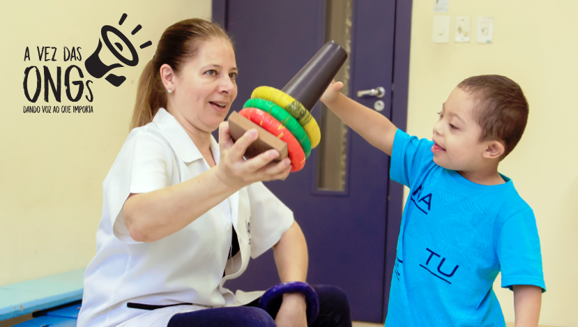 Foto de criança com sindrome de Down brincando de encaixar argolas coloridas junto com sua terapeuta