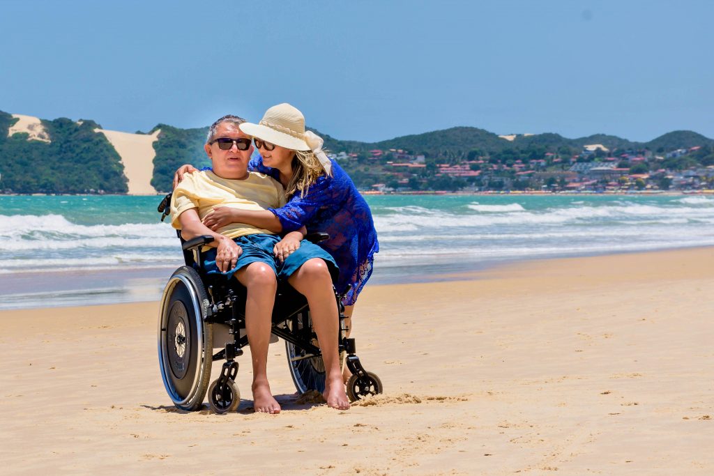 Jose, cadeirante, está na areia da praia e é abraçado por sua esposa. Ao fundo, o mar.
