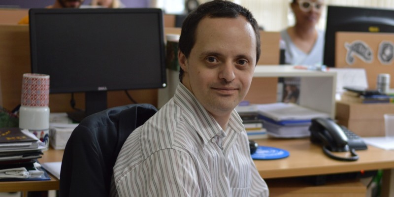 Foto de Edgard, que tem síndrome de Down, posando com sorriso tipo Monalisa. Ele está em sua mesa de trabalho.