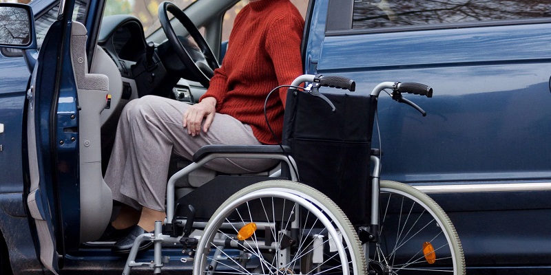 Foto lateral de um carro com a porta do motorista aberta. Uma pessoa tenta sair do carro para sua cadeira de rodas.