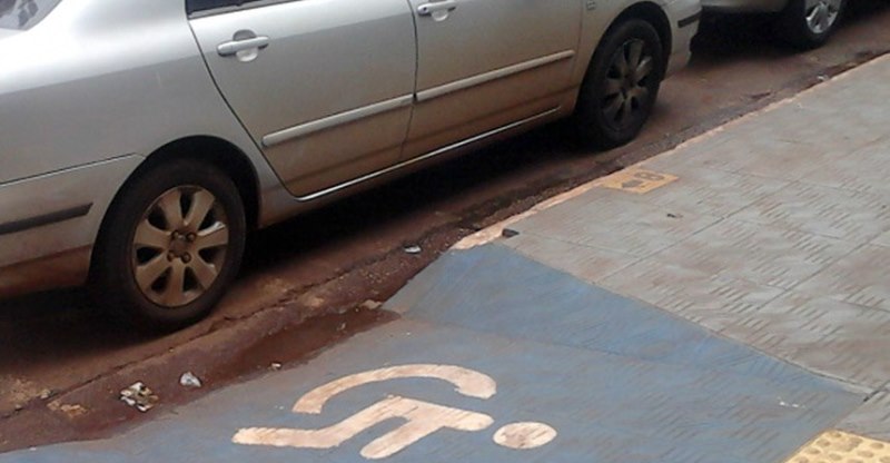 Foto de uma guia rebaixada sinalizada com o símbolo de cadeirante. À sua frente um carro estacionado.