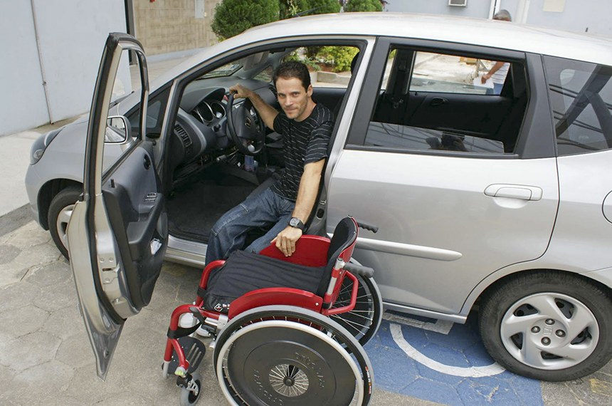 um homem cadeirante está no lugar do motorista em um carro, estacionado em cima de uma vaga reservada. Sua cadeira de rodas está fora do carro ao seu lado.