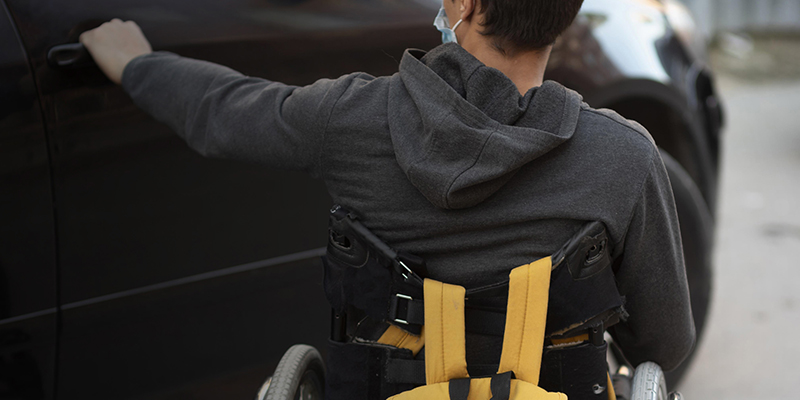 Foto de um rapaz em sua cadeira de rodas de costas prestes a abrir a porta de um carro cinza. 