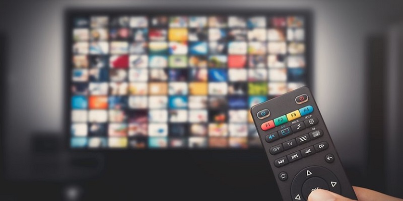 imagem de uma mão segurando o controle remoto apontando para uma televisão desfocada, onde aparece um catálogo de filmes.