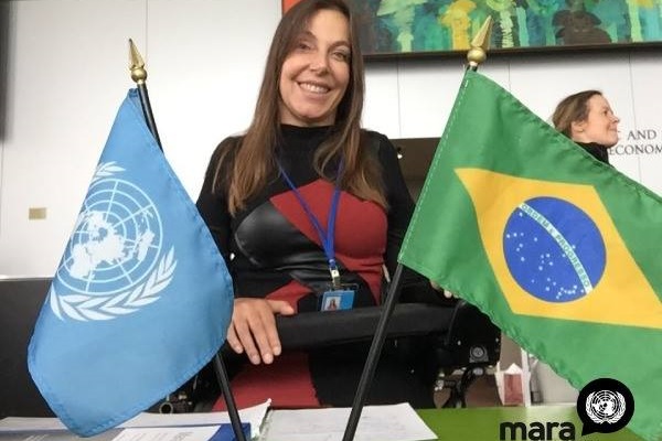 Mara, atrás de uma mesa, sorri entre duas bandeiras colocadas sobre ela. A da esquerda, a da ONU, e a da direita, do Brasil.