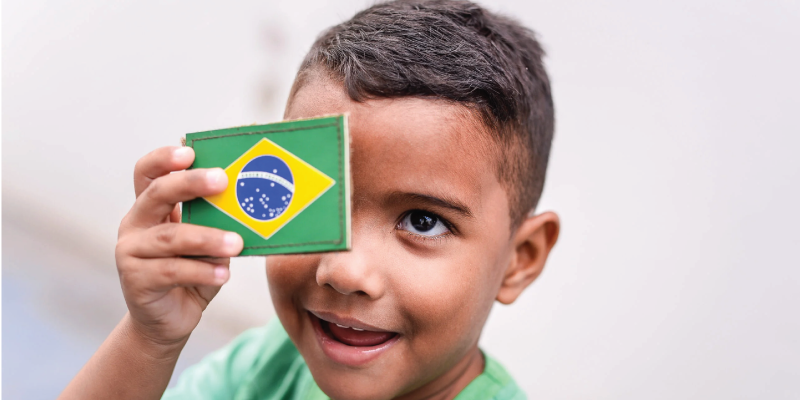 Foto próxima do rosto de uma criança que cobre seu olho esquerdo com uma bandeira pequena do Brasil
