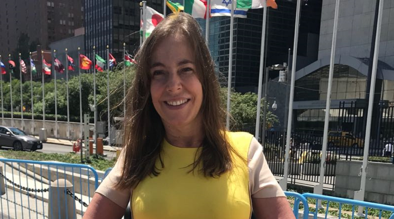 Foto de Mara em frente ao prédio da ONU em Nova Iorque. Atrás dela estão bandeiras de diversos países.