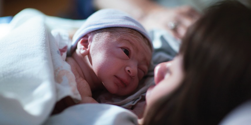 Foto de uma mãe deitada com o seu bebê recém nascido sobre o colo, ele olha para ela