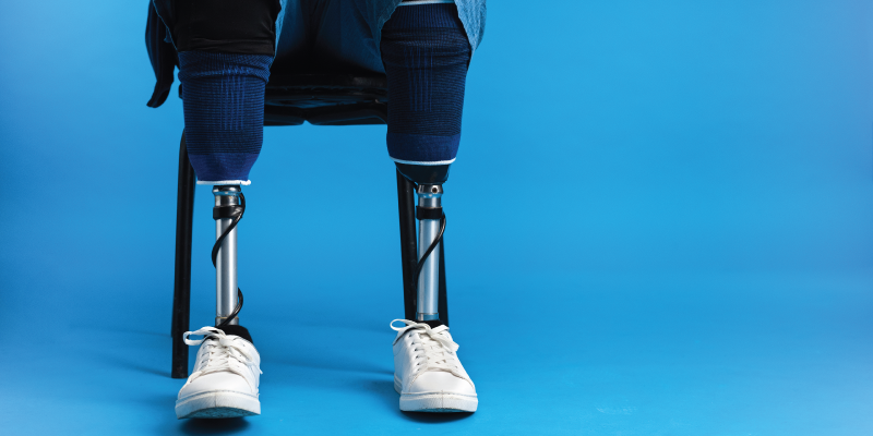 Um foto de uma pessoa com ambas as pernas amputadas sentada em uma cadeira, no lugar duas próteses de metal. O fundo é composto por uma cor azul céu.