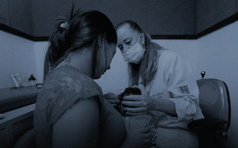 Uma foto com filtro azul escuro. Na foto há uma agente de saúde atendendo uma mãe com um bebê no seu colo