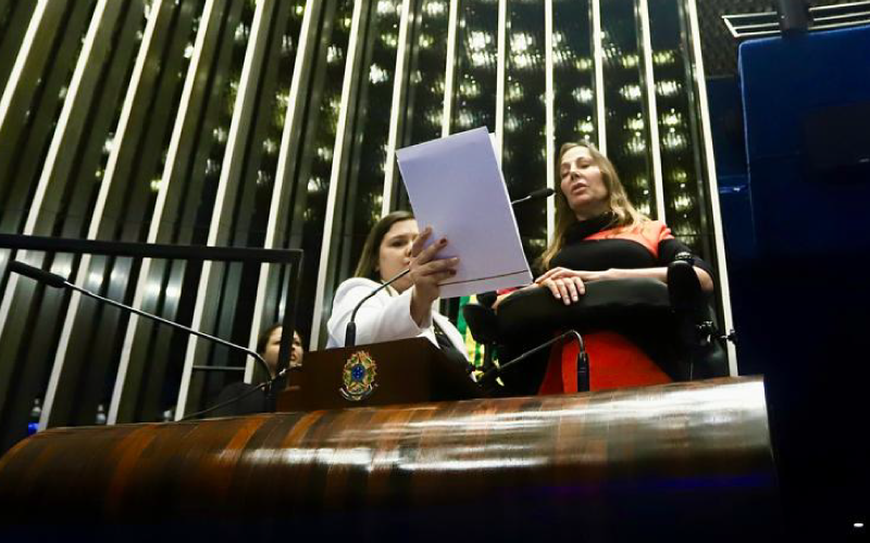 Mara estaem é na sua cadeira na tribuna do senado. Ao lado dela uma assessora segura uma folha de papel em sua frente.