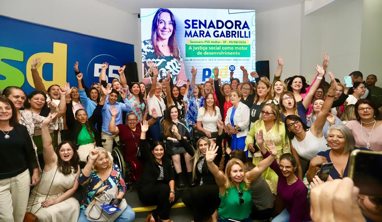 Na imagem Mara está no centro no palco e em sua volta estão várias outras mulheres que participaram no evento. Ao fundo um telão com a arte do convite do evento.