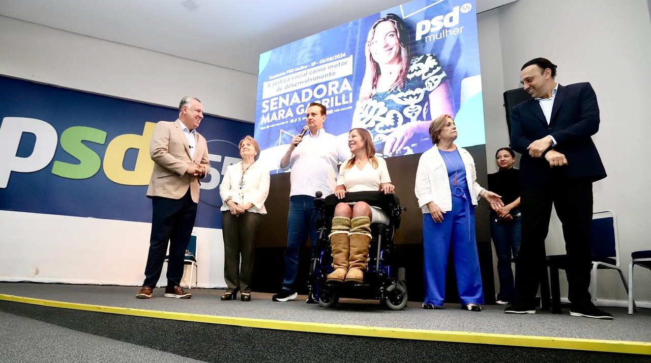 Na foto Mara está ao lado do presidente do PSD Gilberto Kassab que discursa no microfone. Outras autoridades do partido estão no entorno.