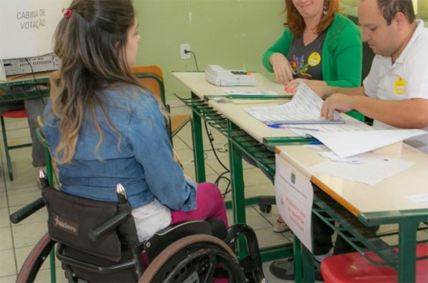 Na imagem uma mulher cadeirante vai até os mesários para poder votar